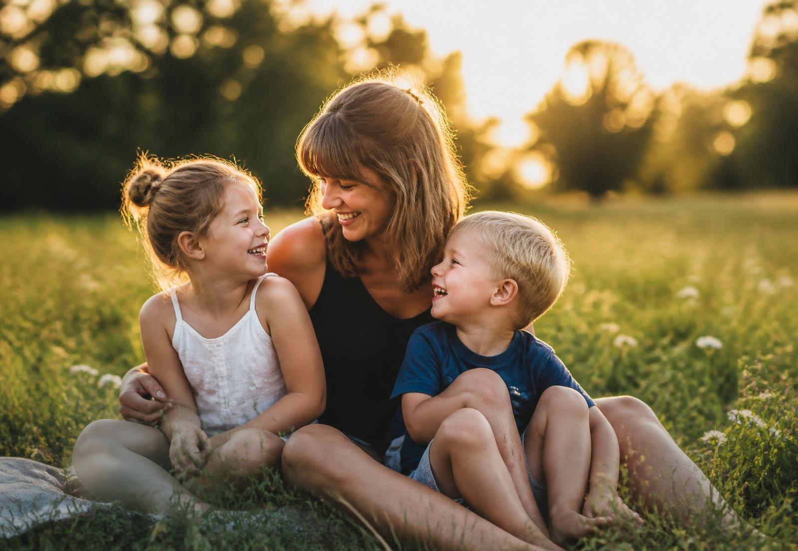 Mutter sitzt lachend mit ihren beiden Kindern auf einer sonnigen Wiese